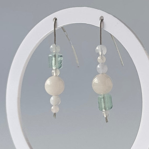 A pair of dangling earrings displayed on a white circular stand against a soft gray studio background. The earrings have silver fishhook wires with small white beads, a pale green faceted bead, and a larger milky white round bead.