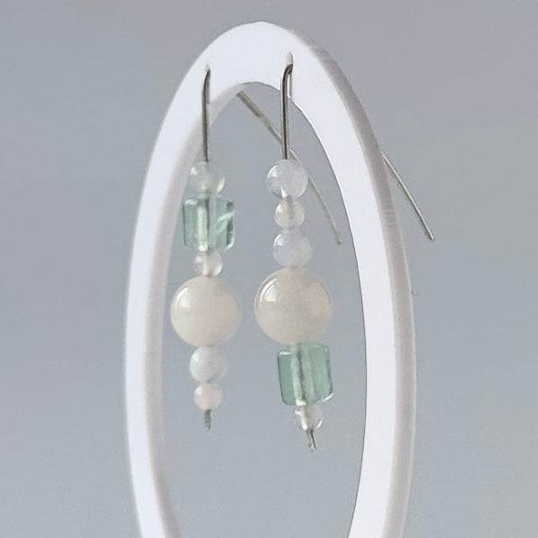 A pair of dangling beaded earrings displayed on a white oval stand against a soft light gray studio background. The earrings feature white pearl-like round beads, small translucent pale green beads, and silver fishhook wires.