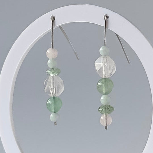 A pair of beaded dangle earrings displayed on a white circular stand against a light gray studio background. The earrings feature clear faceted beads, pale green round beads, and small white beads on silver hooks.