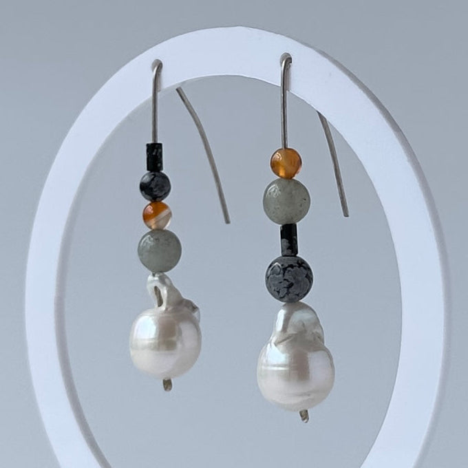 A pair of beaded drop earrings displayed on a white circular stand against a light grey background. Each earring has a silver fishhook wire, small dark and gray beads, a translucent amber bead, a round grey stone bead, and a large white baroque pearl drop.