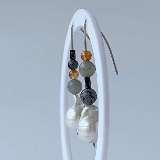 A pair of beaded drop earrings displayed on a white oval stand against a light grey studio background. The earrings feature white baroque pearl drops with small beads in black, grey and orange-brown.
