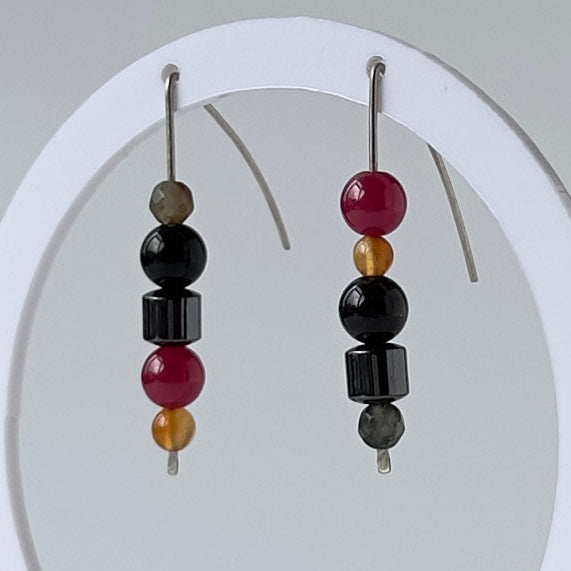 A pair of beaded drop earrings displayed on a white circular stand against a light gray background. The earrings feature black and magenta beads with small gold and gray accents.