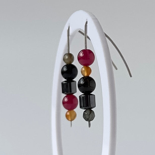 A pair of beaded drop earrings displayed on a white oval stand against a light gray background. The earrings feature alternating black and red beads with small metallic gold and silver spacer beads.