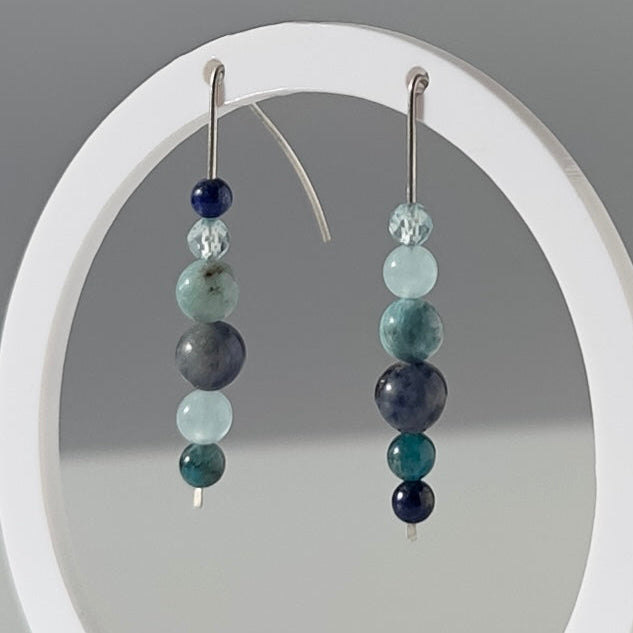 A pair of beaded dangle earrings displayed on a white circular stand against a light gray studio background. The earrings feature small polished beads in dark navy, teal, green, and gray tones with silver hooks.