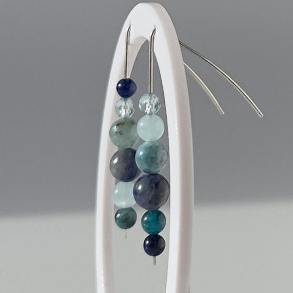 A pair of beaded drop earrings displayed on a white oval stand against a light gray background. The earrings feature multiple horizontal rows of polished beads in shades of navy, teal, green, and gray, suspended from thin silver-toned wires.