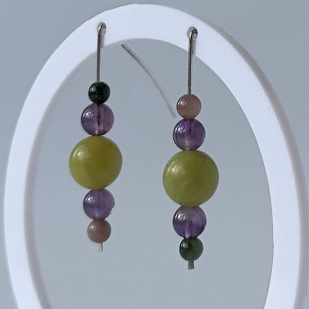 A pair of beaded dangle earrings displayed on a white oval stand against a light gray studio background. The earrings feature large olive green beads, purple beads, and small dark and pinkish beads on silver hooks.