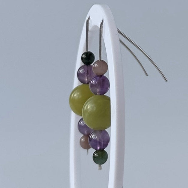 A pair of beaded earrings displayed on a white stand against a light gray background. The earrings feature a cluster of translucent purple beads, a large olive-green bead, a small black bead, and a dark green bead, suspended from thin metal wires.