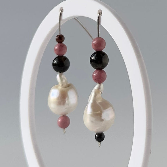 A pair of beaded dangle earrings displayed on a white oval stand against a light gray studio background. The earrings feature cream baroque pearls, black round beads, and dusty pink round beads.