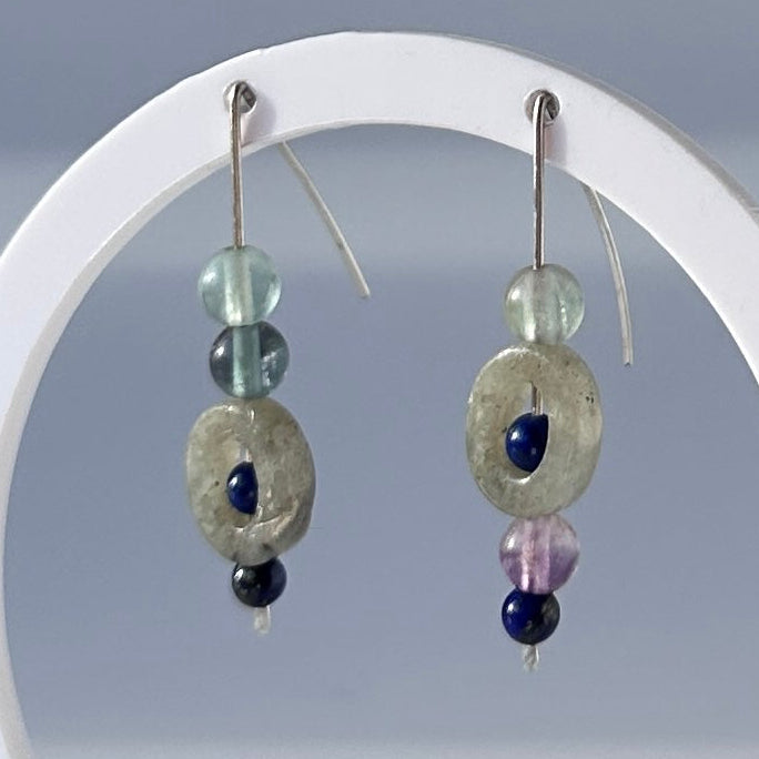 A pair of beaded dangle earrings displayed on a white circular stand against a soft gray background. Each earring has a silver fishhook, a pale translucent bead, a larger iridescent oval bead, a dark blue bead, and a small lavender bead.