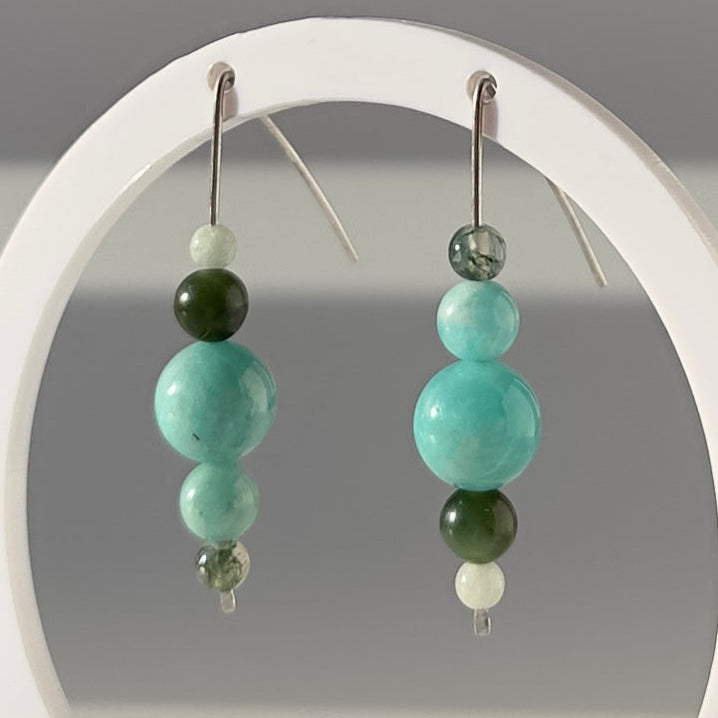 A pair of beaded dangle earrings displayed on a white circular stand against a soft gray background. Each earring features a cluster of beads in light aqua, dark green, white, and a small clear faceted bead, suspended from silver hooks.