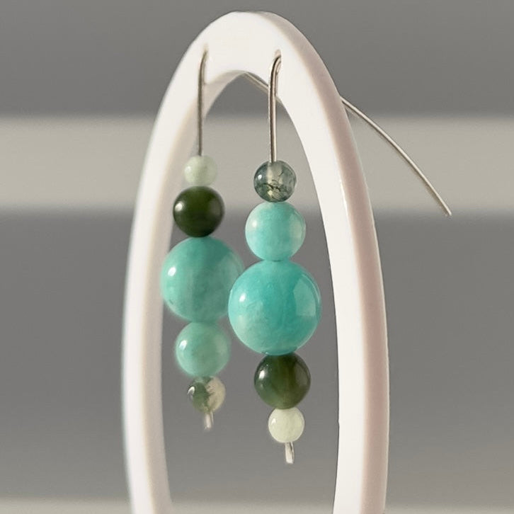 A pair of beaded dangle earrings with light aqua and dark green round beads, hanging from a white oval display stand against a soft gray background.