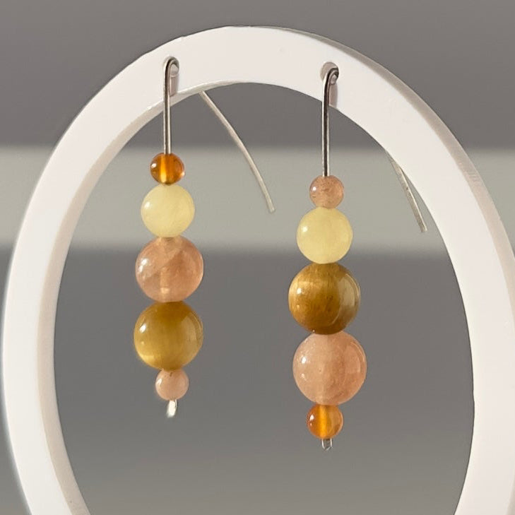A pair of beaded dangle earrings displayed on a white oval stand against a soft gray background. The earrings feature a mix of pastel yellow, peach, ochre, and light green beads with silver-toned hooks.