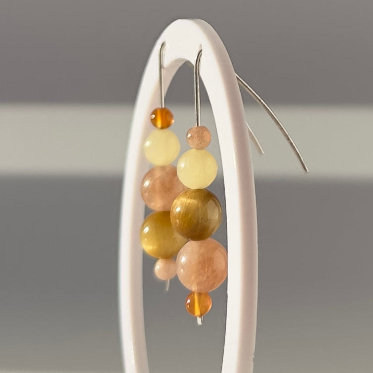 A pair of beaded dangle earrings displayed on a white ring stand against a soft gray background. The earrings feature a mix of translucent and opaque beads in pale yellow, peach, and metallic gold, suspended from thin silver-toned wires.