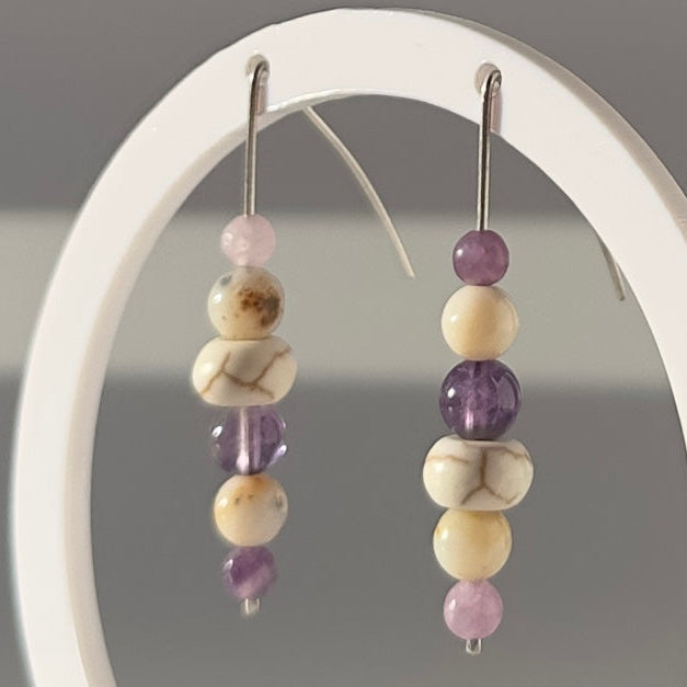 A pair of beaded dangle earrings displayed on a white circular stand against a soft gray background. The earrings feature a mix of lavender purple beads, cream-colored beads, and marbled beige and white beads.