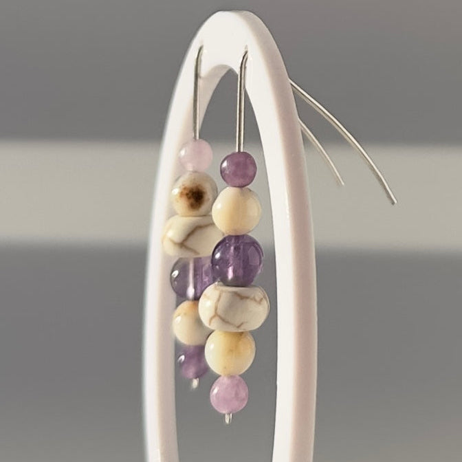 A pair of beaded dangle earrings displayed on a white circular stand against a soft gray background. The earrings feature silver-toned fishhook wires with a cluster of beads including lavender purple, cream, and marbled white beads.
