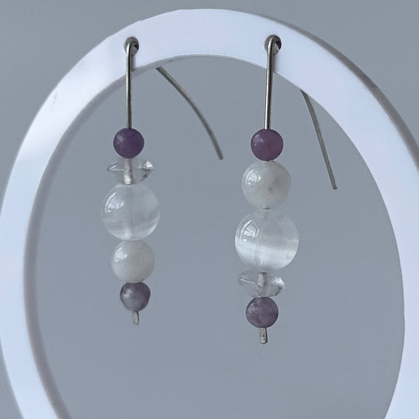 A pair of beaded dangle earrings displayed on a white circular stand against a soft gray studio background. Each earring has a silver fishhook wire, a small dark purple bead, a larger milky white bead, and a smaller translucent white bead with a tiny dark purple bead below.