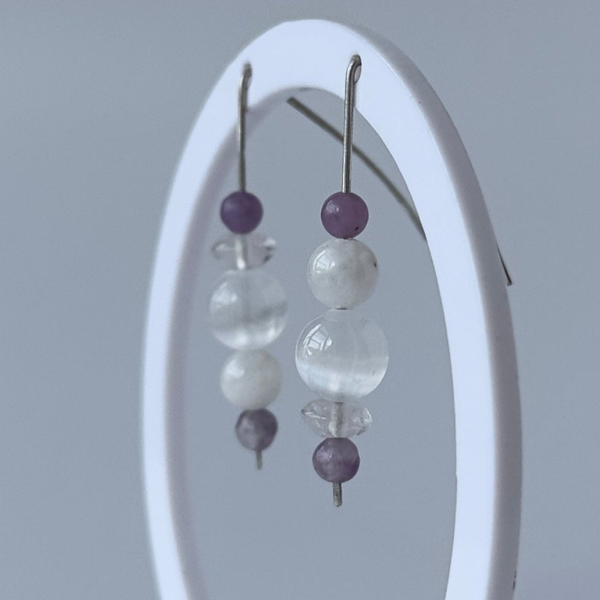 A pair of beaded dangle earrings displayed on a white oval stand against a soft gray studio background. The earrings feature translucent white beads, clear faceted round beads, and small dark purple beads.