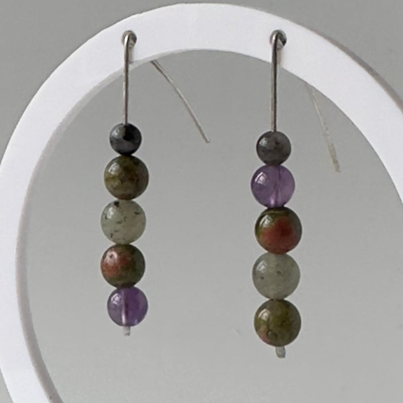A pair of beaded dangle earrings displayed on a white circular stand against a light gray background. The earrings feature multiple polished beads in earthy tones including purple, olive green, brown, and dark gray.