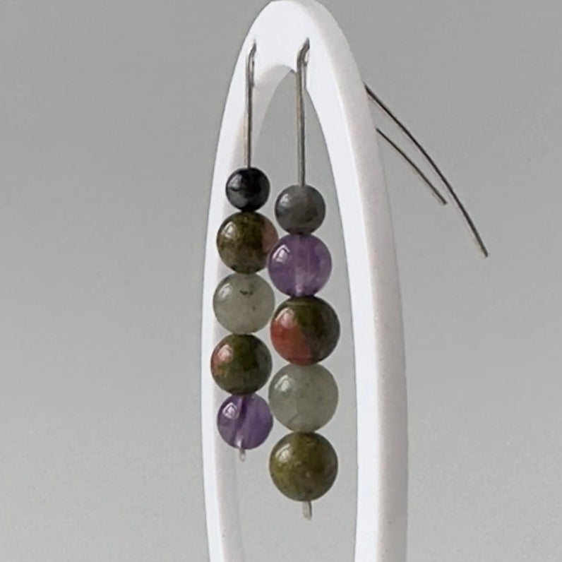 A pair of beaded drop earrings hanging from a white display stand against a plain light gray background. The earrings feature multiple strands of polished beads in dark gray, purple, olive green, and brown tones.