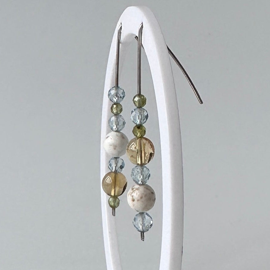 A pair of beaded drop earrings displayed on a white oval stand against a light gray background. The earrings feature clear faceted beads, white beads, and metallic gold and silver beads.