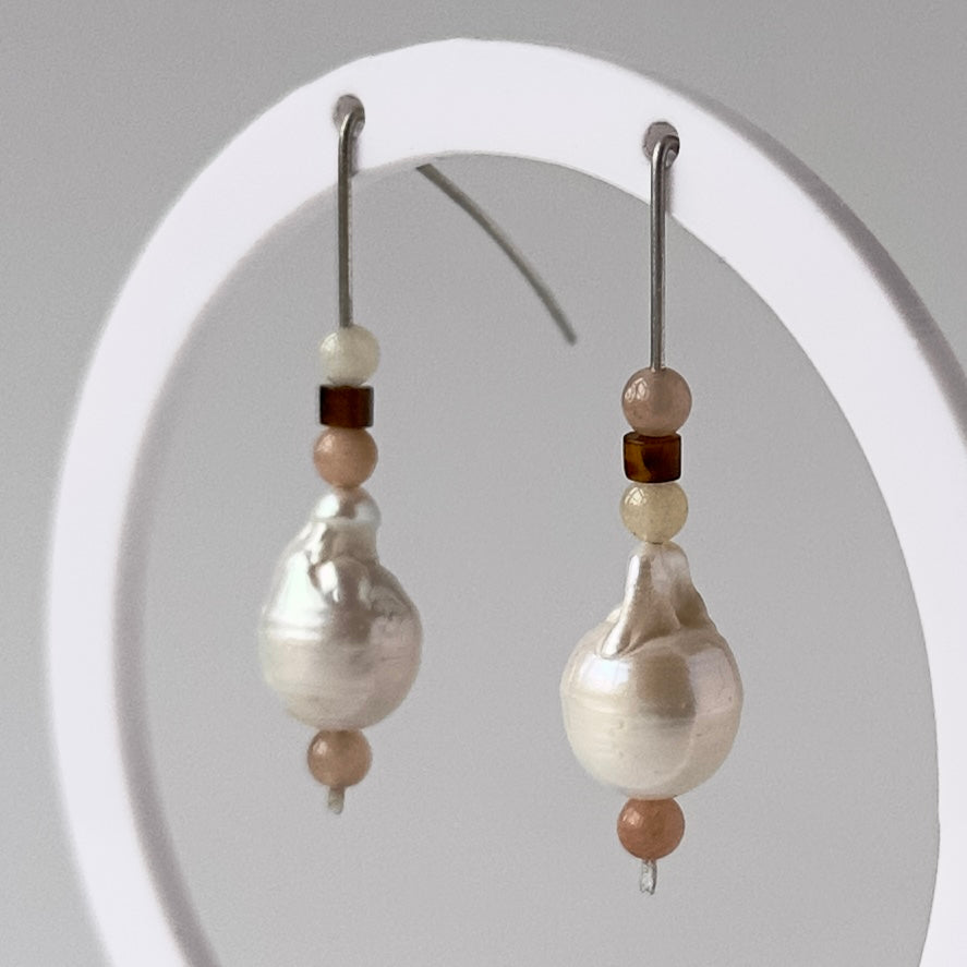 A pair of drop earrings displayed on a white circular stand against a light grey background. Each earring features a large irregular white pearl bead with smaller beige and dark brown beads above it and a silver fishhook wire.