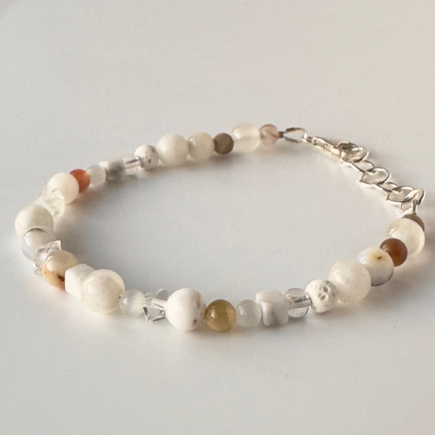 A beaded bracelet with white, cream, and brown stone beads, clear faceted crystal beads, and a silver lobster clasp, displayed on a light gray background.
