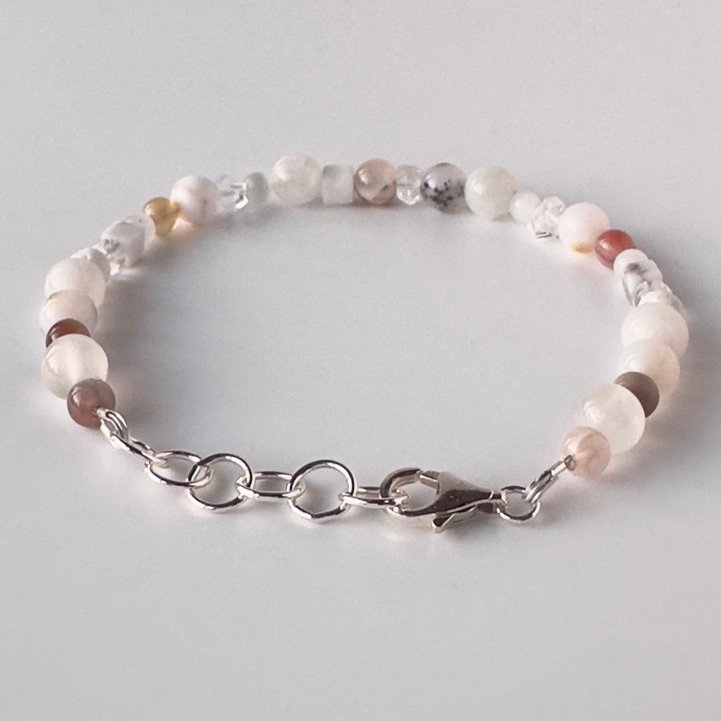 A beaded bracelet with round white, light brown, and clear agate-like beads, connected by a silver metal chain with a lobster clasp, shown on a light grey background.