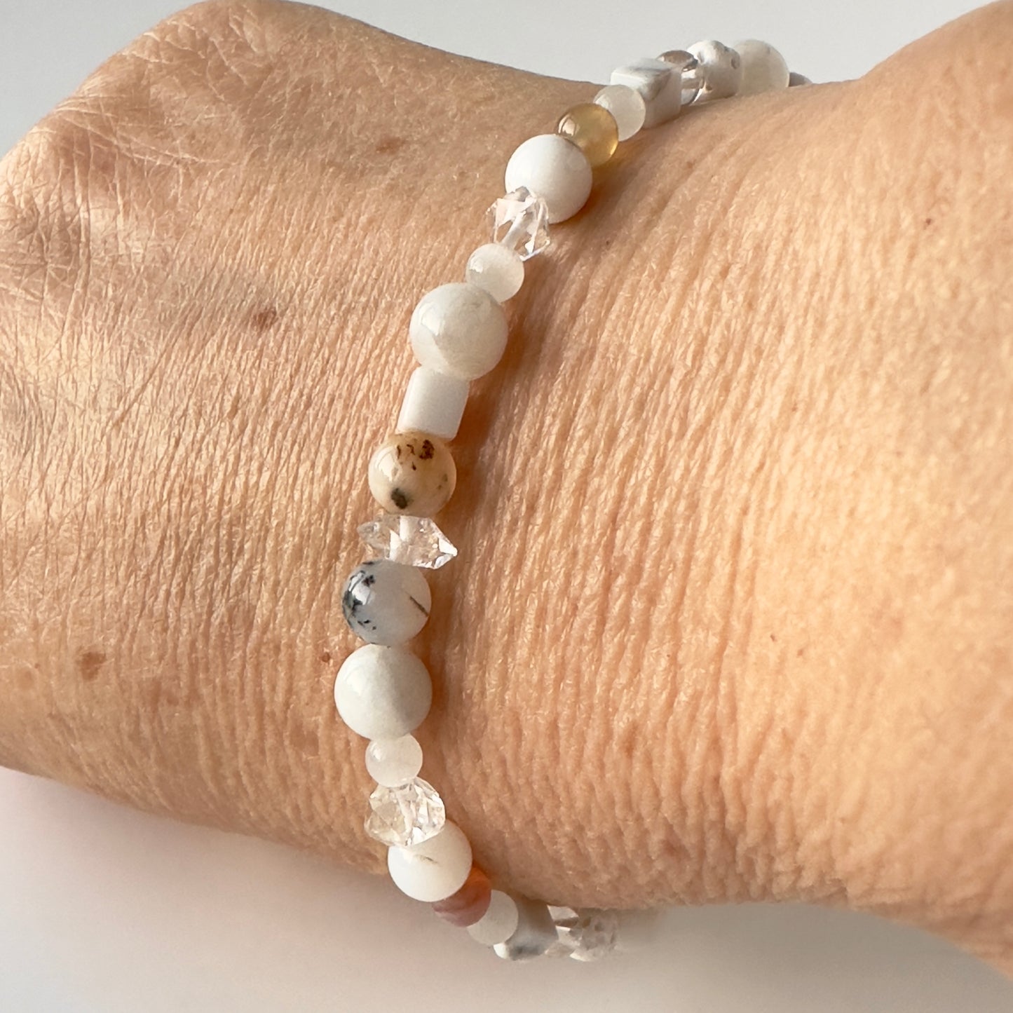 A close-up of a beaded bracelet worn on a person's wrist. The bracelet features matte white beads, faceted clear crystal beads, and a few small tan and speckled brown beads.