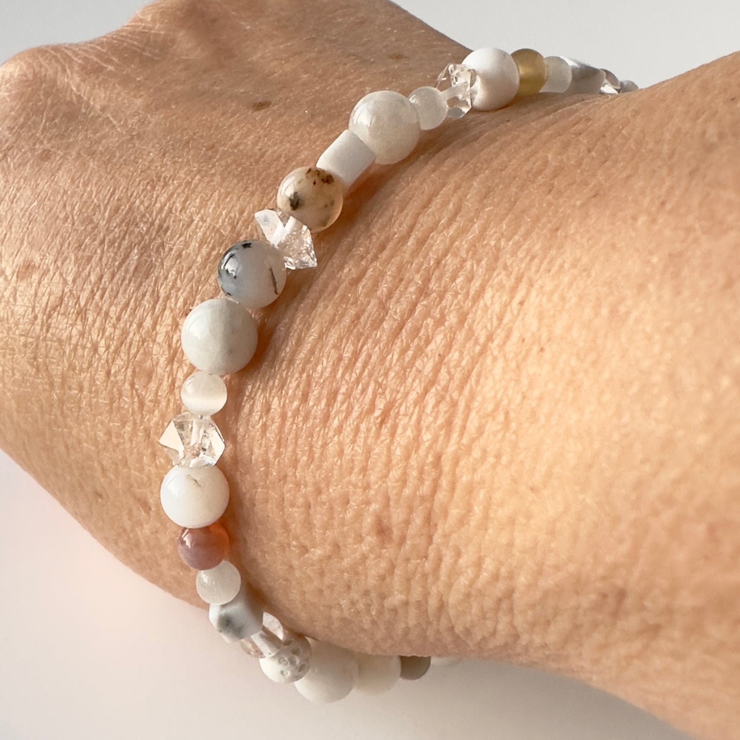 A close-up of a beaded bracelet worn on a person's wrist. The bracelet features matte white and cream beads, faceted clear crystal beads, and a few marbled brown and grey beads.