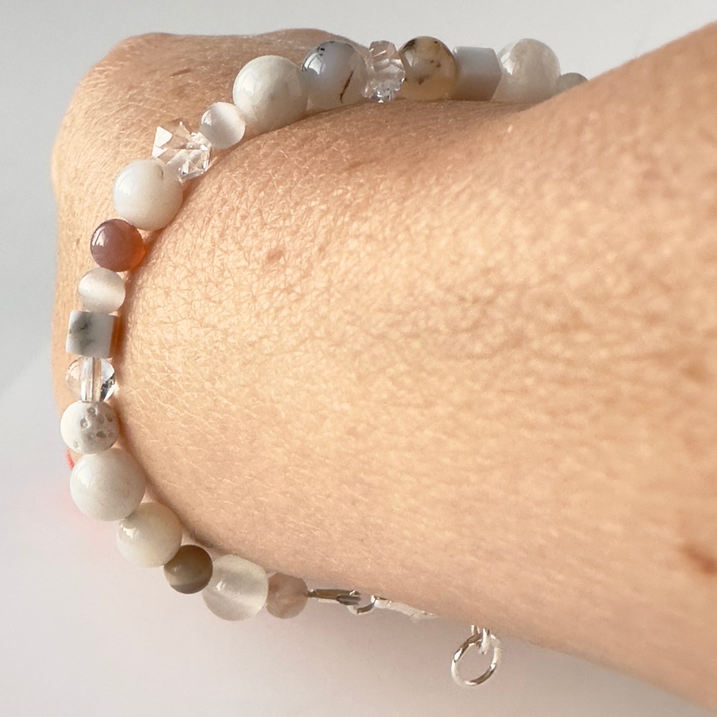 A close-up of a beaded bracelet worn on a person's wrist. The bracelet features matte white and grey beads, faceted clear crystal beads, and a few brown marbled beads, with a silver clasp visible at the bottom.
