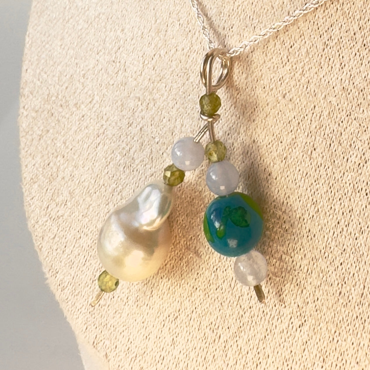 A close-up of a beaded necklace displayed on a cream fabric jewelry bust. The necklace features a large baroque pearl, a round blue and green bead, small faceted green crystals and small translucent beads on a silver-toned chain.
