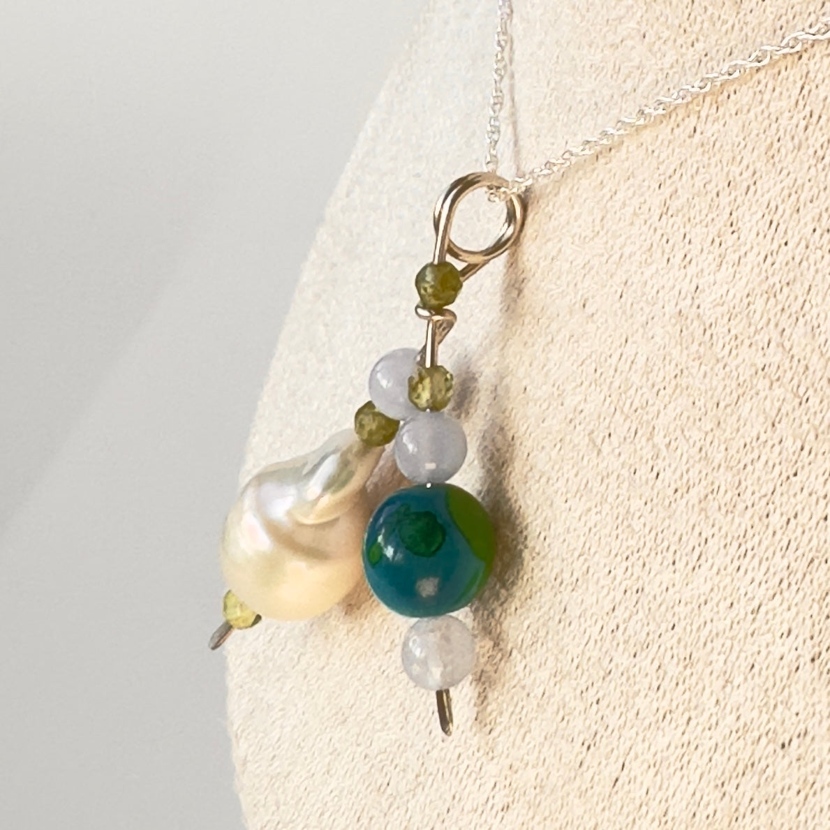 A close-up of a beaded pendant necklace displayed on a cream fabric jewelry bust. The necklace features a silver chain, a cluster of beads including a large baroque pearl, a round green-and-blue bead, green faceted crystals and small translucent white beads.