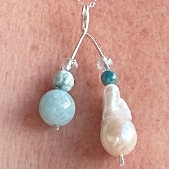 A close-up of a silver necklace worn on a person's chest, featuring a pendant with a silver double-hanging design, a light blue round bead, a small clear bead, a dark blue bead, and a white baroque pearl.