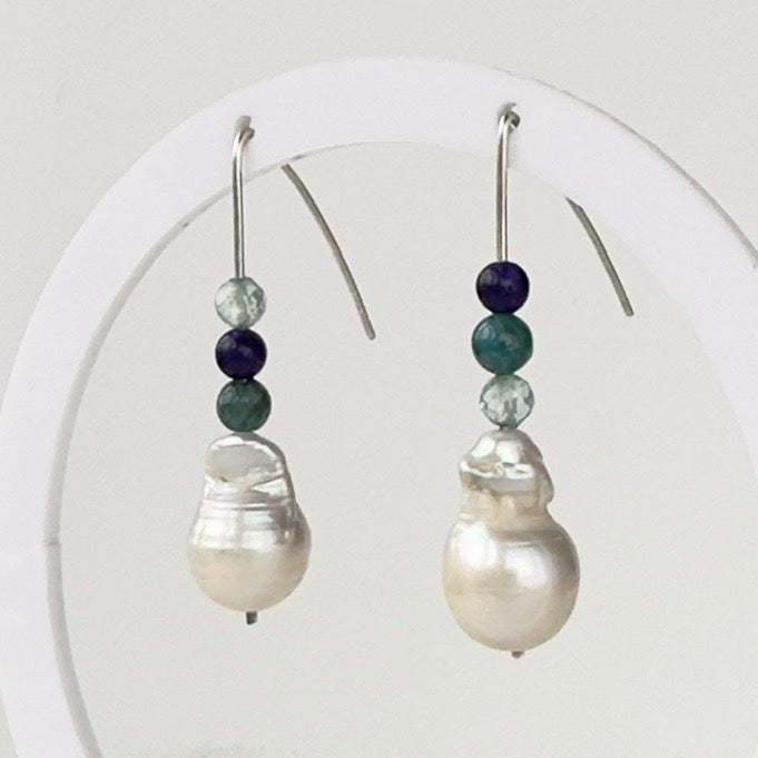 A pair of silver drop earrings displayed on a white circular stand. Each earring has a large irregular white baroque pearl drop with three small beads above it in dark blue, teal, and light blue crystal..