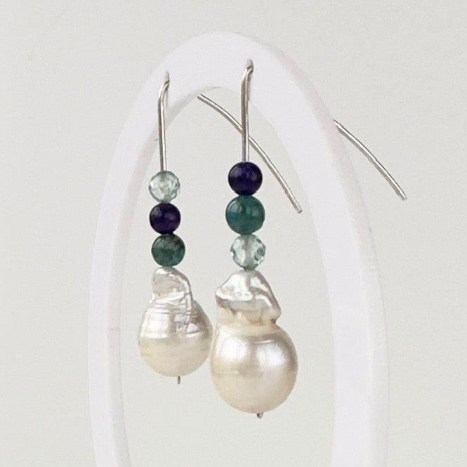 A pair of silver fishhook earrings with baroque pearl drops and teal, light blue and dark blue beads, displayed on a white arch stand.