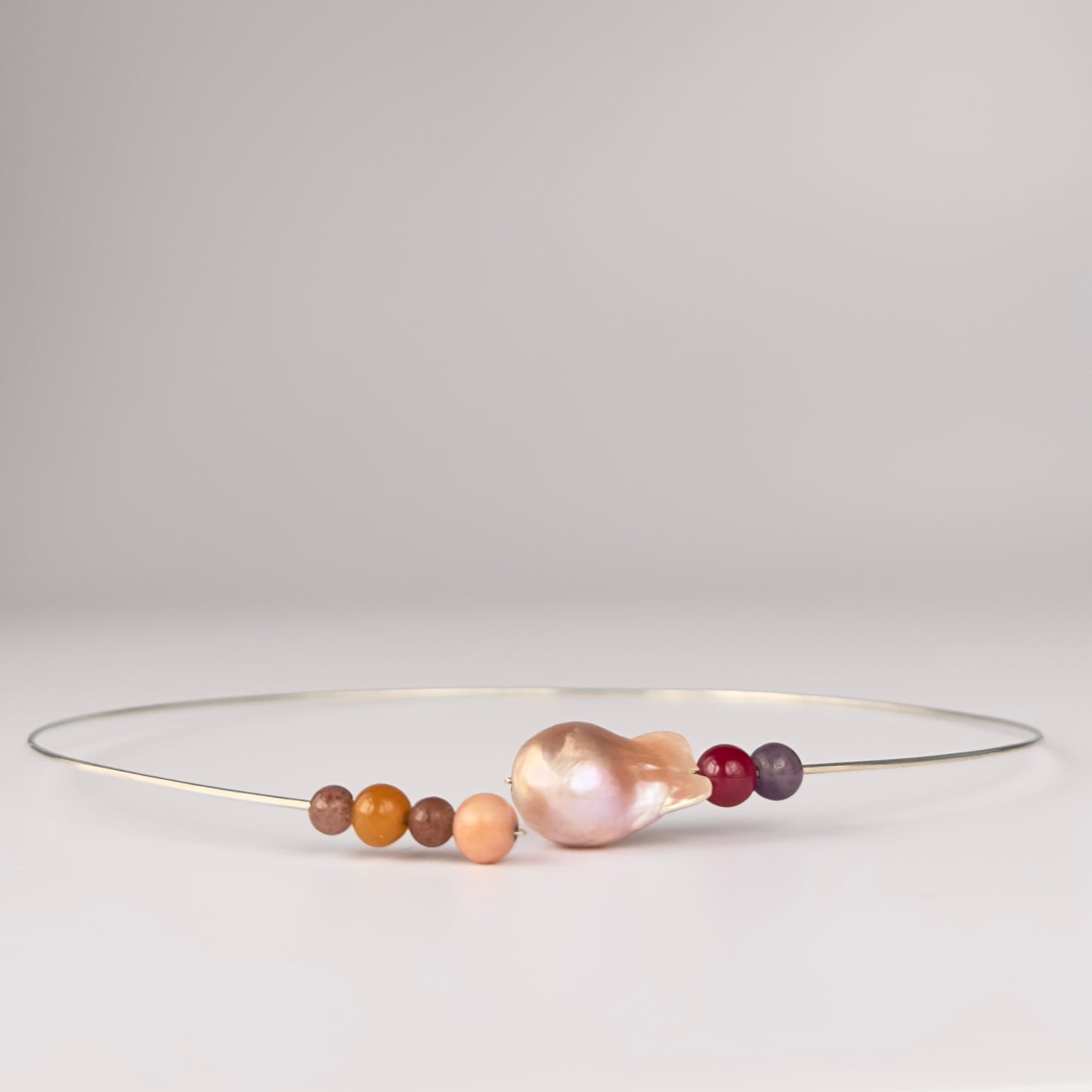 A delicate silver wire bangle style necklace featuring a large, irregular baroque pearl in a soft pinkish hue, accented by small smooth beads in orange, brown, peach, and deep red, set against a neutral light grey background.