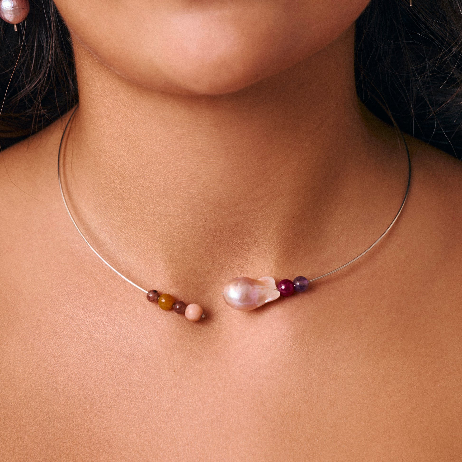 A close-up of a delicate silver wire necklace worn on a model's neck, featuring a large baroque pearl and small colourful beads.