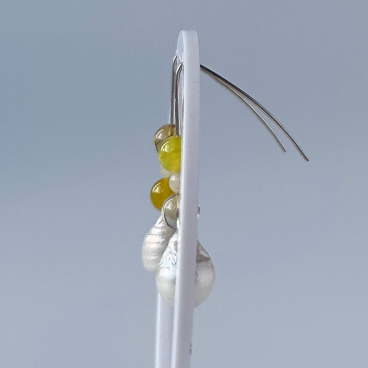 A pair of dangle earrings displayed on a white stand against a light grey background. The earrings feature yellow beads, white baroque pearls, and silver handmade hooks.