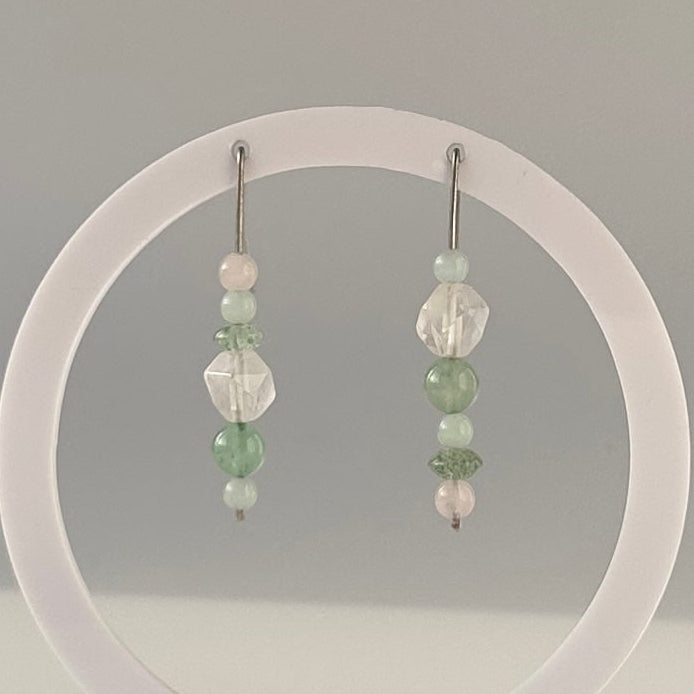 A pair of drop earrings with pale green and clear crystals displayed on a clear circular stand on a white surface, photographed in a softly lit indoor setting.