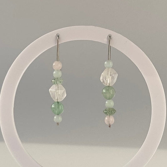 A pair of drop earrings with pale green and clear crystals displayed on a clear circular stand on a white surface, photographed in a softly lit indoor setting.