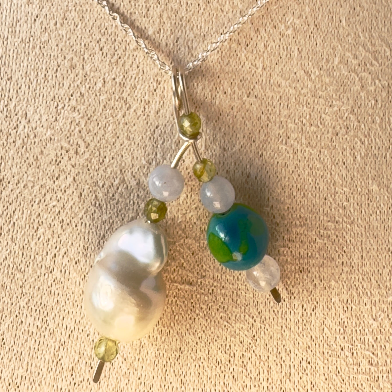 A close-up of a delicate necklace with a silver chain and a multi-bead pendant featuring a large white baroque pearl, a blue and green marbled bead, small translucent blue beads, and ssmall faceted green crystals displayed on a textured beige fabric background.