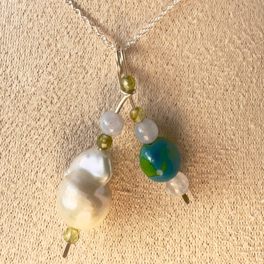 A close-up of a delicate necklace with a silver chain and a multi-bead pendant featuring a large white baroque pearl, a blue and green marbled bead, small translucent blue beads, and ssmall faceted green crystals displayed on a textured beige fabric background.