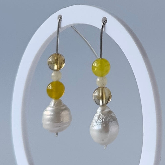 A pair of beaded drop earrings displayed on a white oval stand against a soft light grey background. The earrings feature silver hooks, yellow beads, and baroque pearls