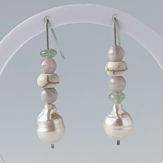 A pair of handmade beaded drop earrings displayed on a white circular stand against a light grey background. The earrings feature a vertical stack of beads: a light green disc-shaped bead, pale pink round beads, a white bead with dark veining, and a large lustrous white baroque pearl at the bottom.