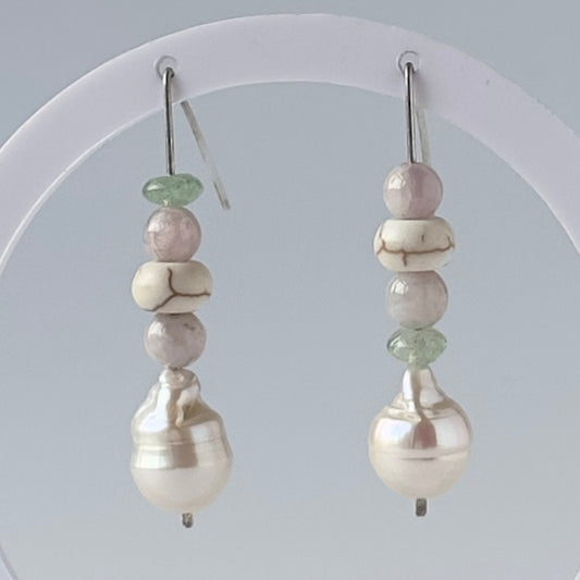 A pair of handmade beaded drop earrings displayed on a white circular stand against a light grey background. The earrings feature a vertical stack of beads: a light green disc-shaped bead, pale pink round beads, a white bead with dark veining, and a large lustrous white baroque pearl at the bottom.